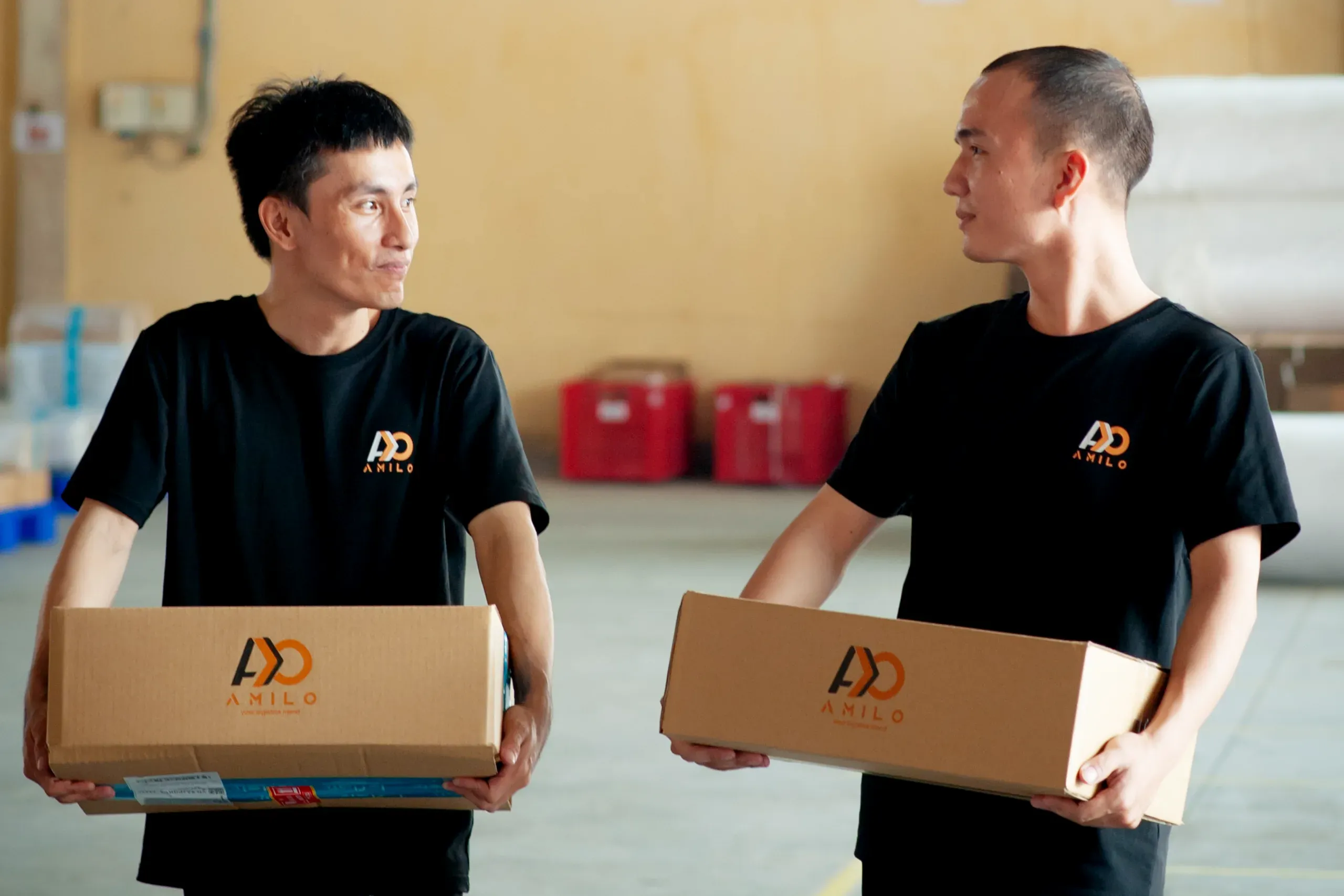 Two Amilo warehouse team members in branded black t-shirts smiling at each other while each carries an Amilo-branded box.